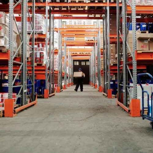 A worker carrying a box in a well-organized warehouse storage aisle.